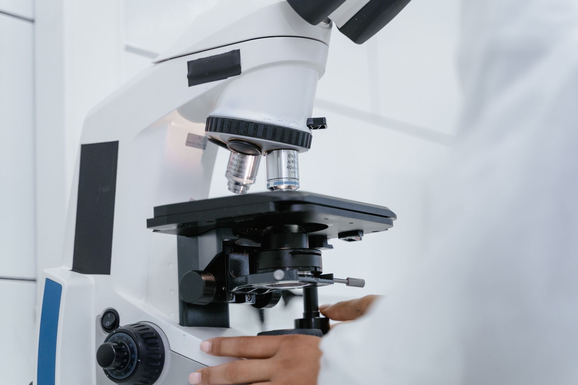 Gros plan sur les objectifs et la platine d’un microscope optique manipulé par les mains d’une personne en blouse blanche.