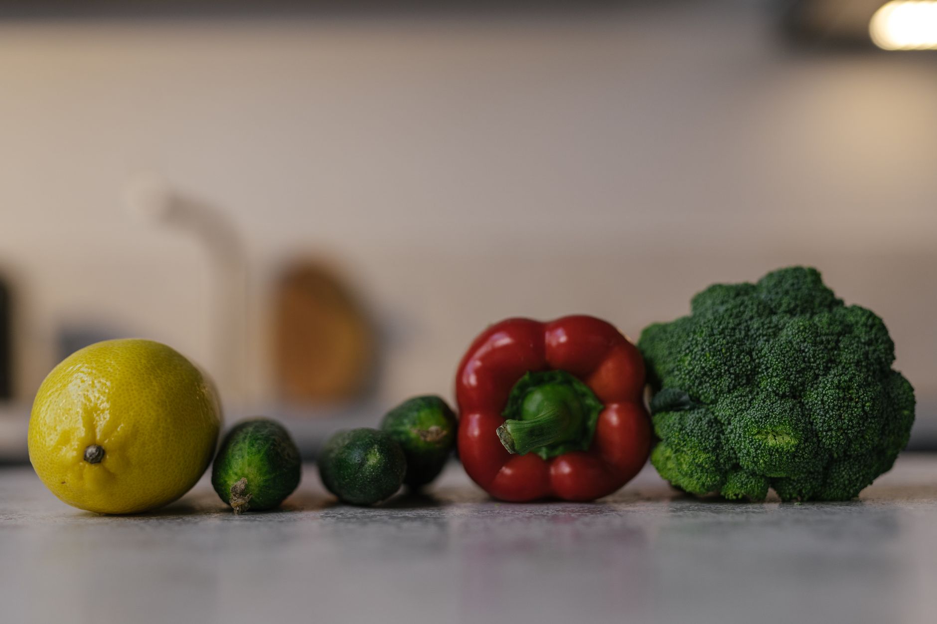 Un citron, trois petits concombres, un poivron rouge et un brocoli alignés sur un plan de travail gris dans une cuisine floue.